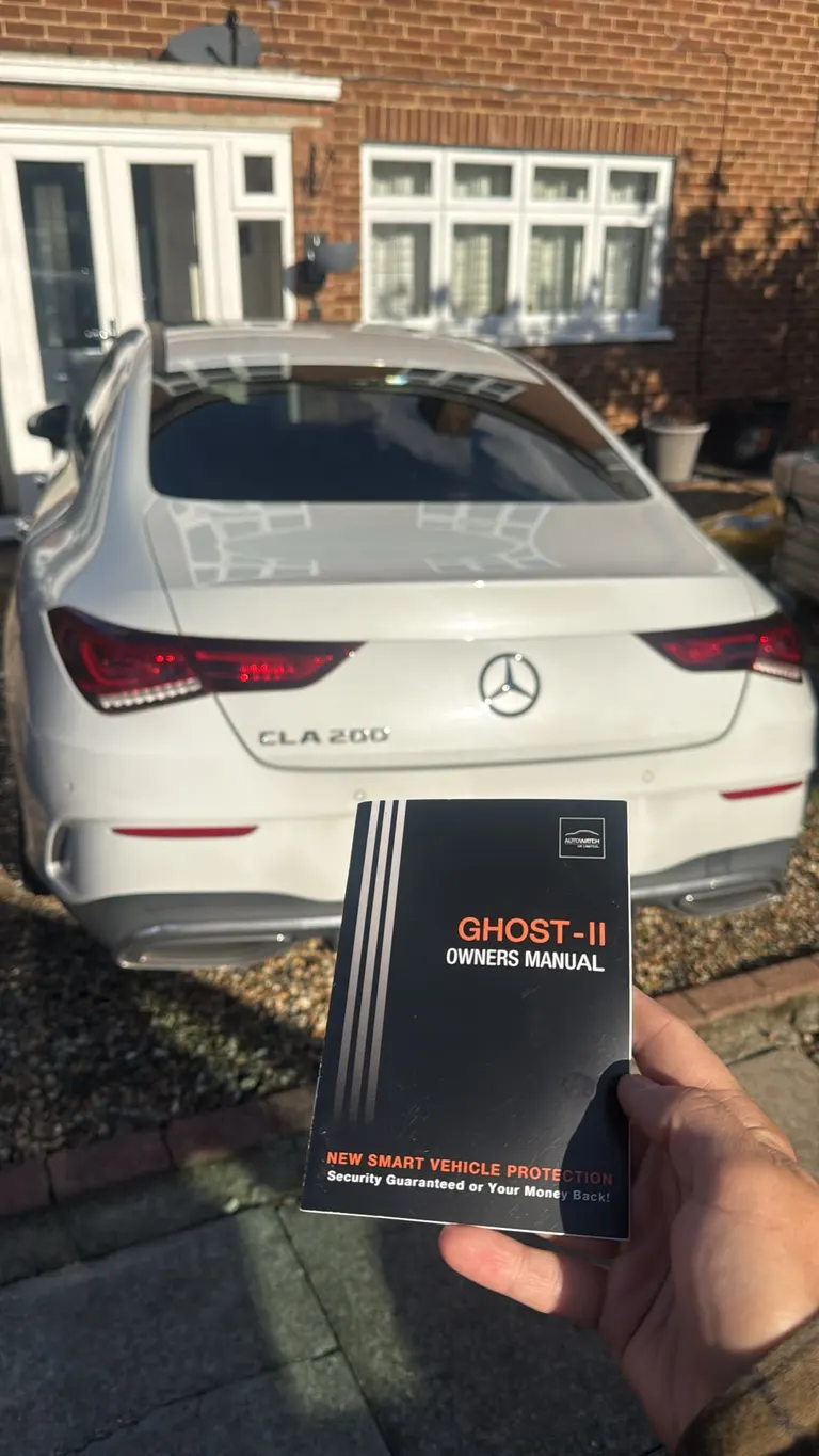 Autowatch Ghost-II owner's manual held in front of a white Mercedes CLA 200 on a driveway in Esher after the immobiliser was fitted