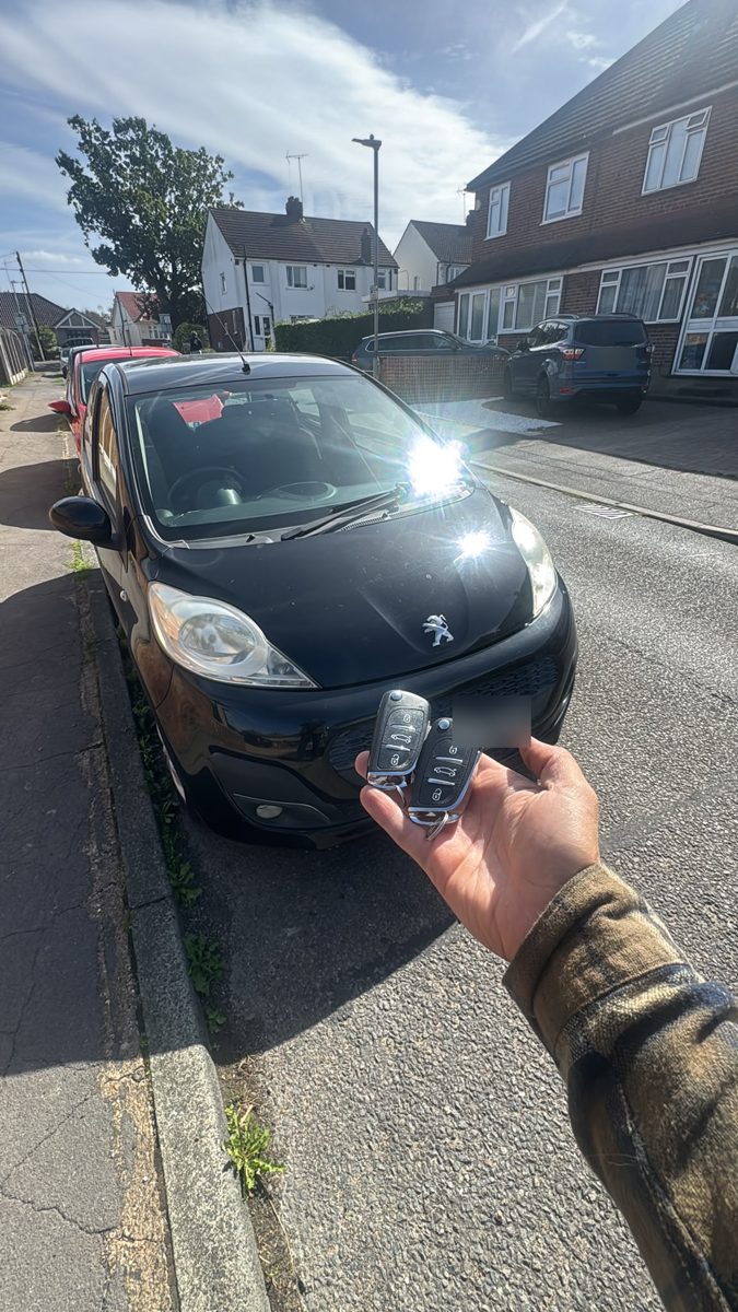 Two new Peugeot flip keys held in front of a black Peugeot 107 in Billericay after an all-keys-lost job
